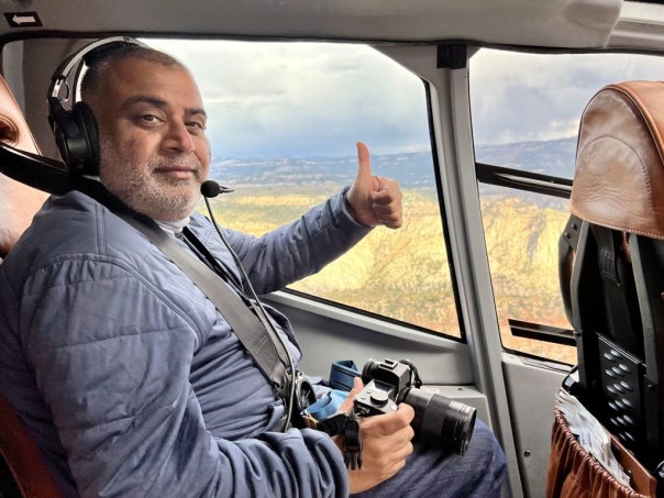 Smiling man wearing a headset seated in a helicopter cockpit, giving a thumbs up while holding a camera. Scenic landscape outside the window.
