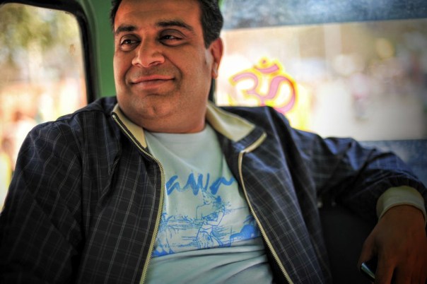Smiling man in a blue graphic T‑shirt and dark plaid jacket sits in a vehicle, looking off to the side.